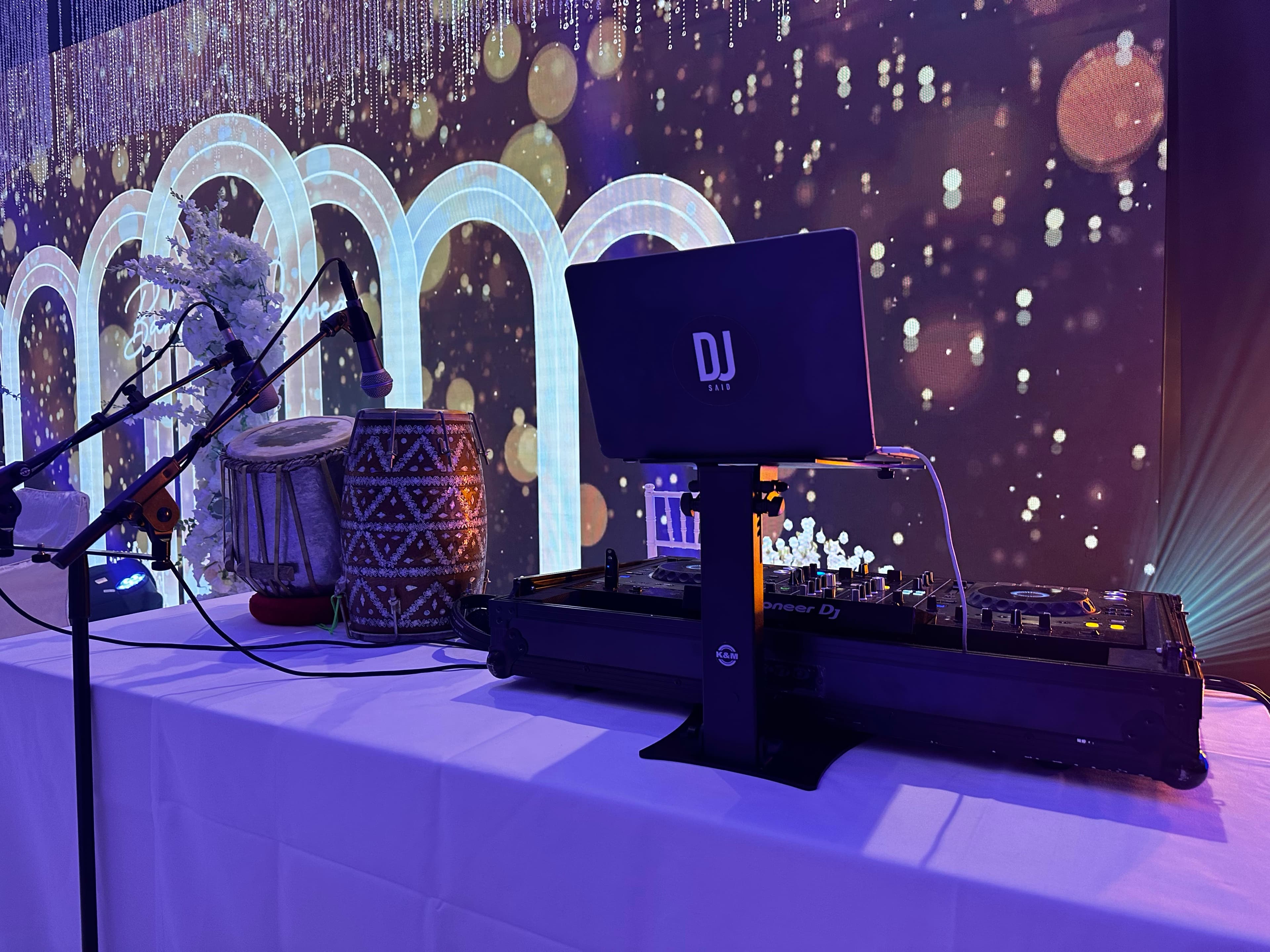 Aufgebautes DJ-Setup von DJ Said im Festsaal: Pioneer-Controller, Laptop, Funkmikrofon und afghanische Dhol vor einer beleuchteten LED-Wand